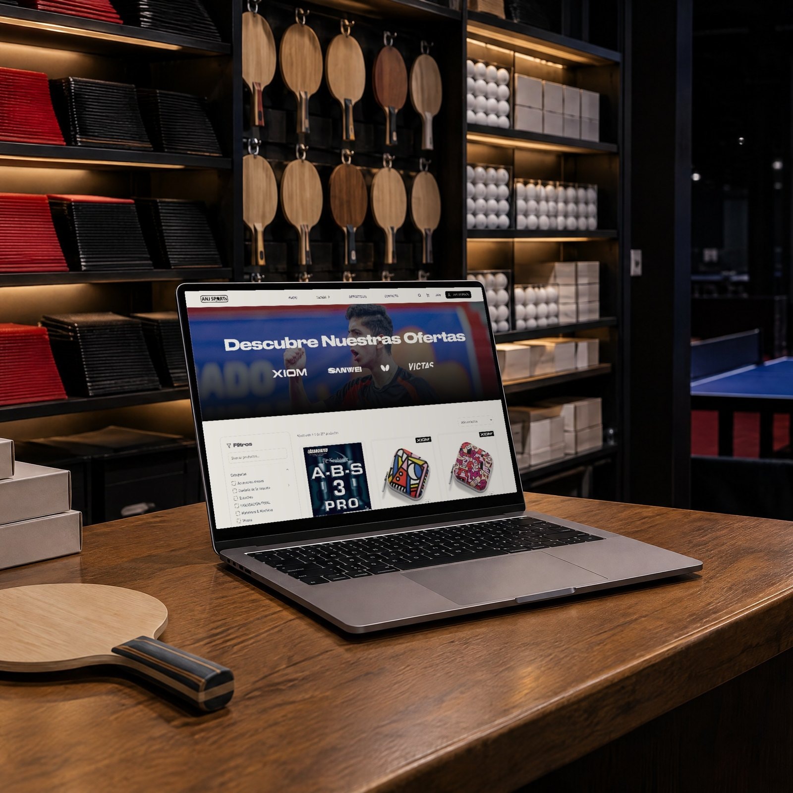 Una laptop en el mostrador de una tienda de tenis de mesa, con el catálogo de anjsports.com y una pared de gomas y paletas detrás