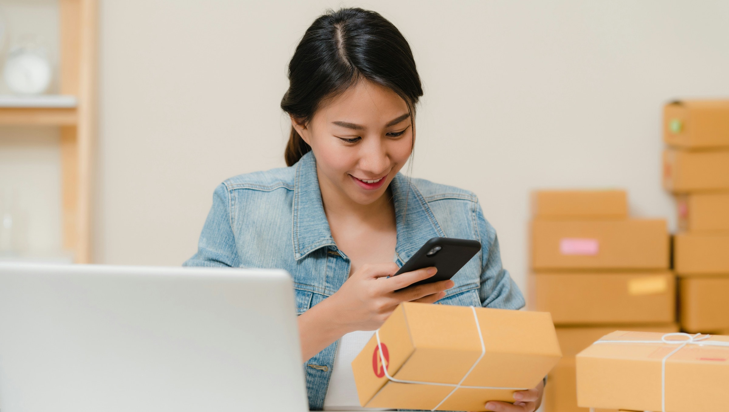 Una emprendedora revisa un pedido con el celular, entre la laptop y una pila de cajas por despachar