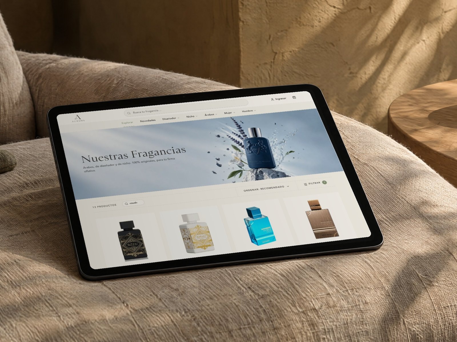 Una tableta sobre un sillón de lino muestra la búsqueda «oud» en aurore.com.pe: trece fragancias de casas distintas
